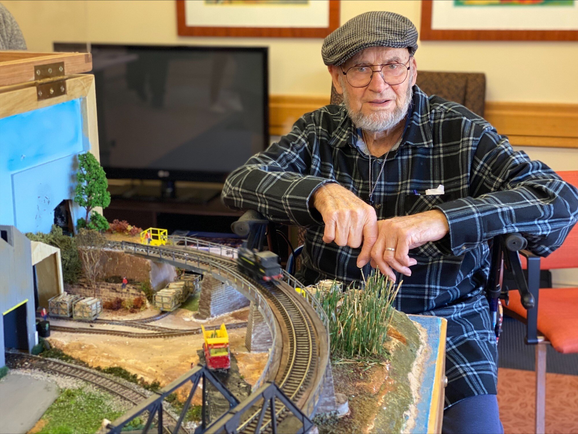 Man with grey beard and cap sits in chair beside model train set