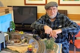 Man with grey beard and cap sits in chair beside model train set