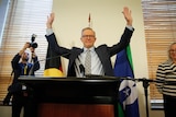 Albanese at the lectern has both hands in the air, waving to the room, as photographers take pictures