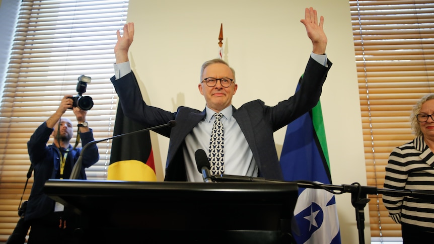 Albanese at the lectern has both hands in the air, waving to the room, as photographers take pictures