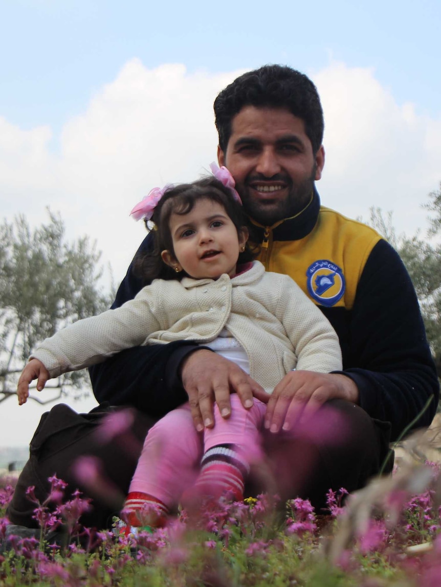 A man sits among flowers with a little girl with pigtails in his lap.