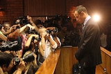 Man in a suit stands for his bail hearing in front of a crowd of photograpers.