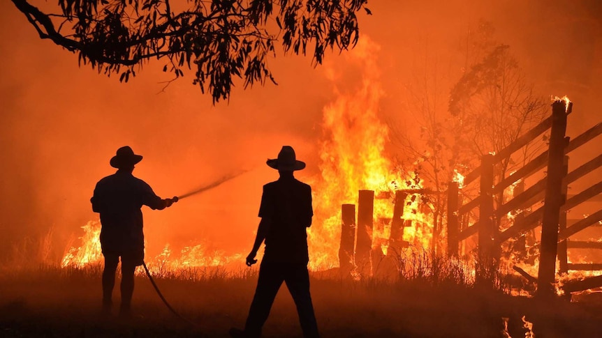 Silhouettes of two people in broad-brimmed hats, one holding a hose, against bright red and orange flames