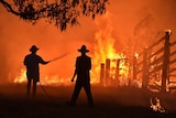 Silhouettes of two people in broad-brimmed hats, one holding a hose, against bright red and orange flames