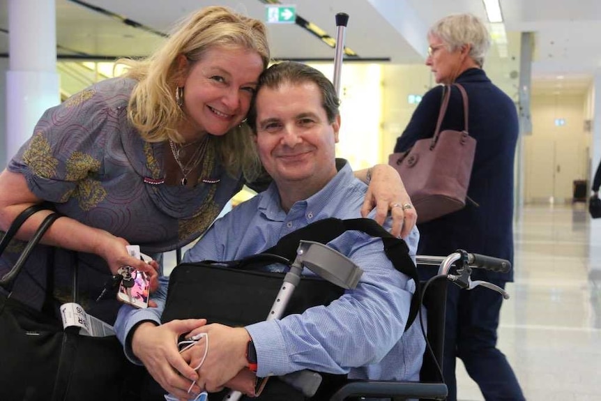 Desiree and Anthony smile happily at the camera, he in a wheelchair.