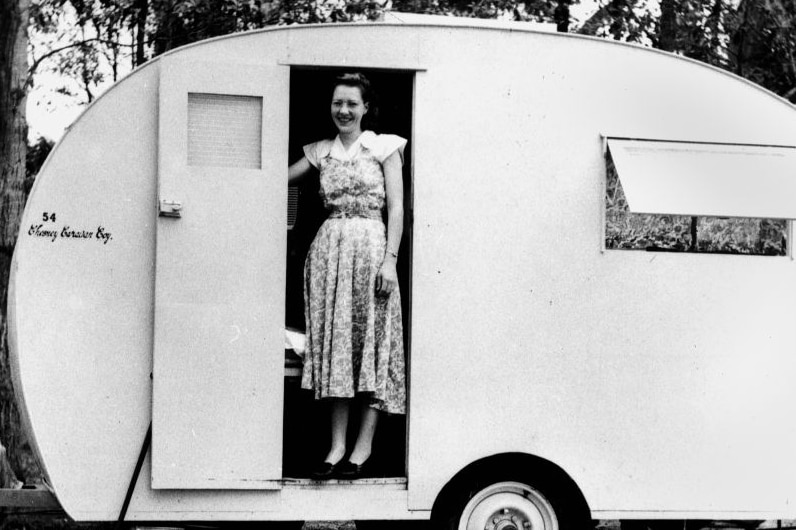 Black and white photo of a woman in 1952 wearing a knee length dress, standing in the door of a Chesney caravan.