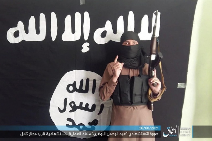 A masked Islamic State fighter poses in front of an IS flag with a rifle.