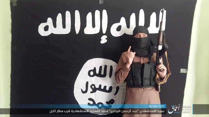 A masked Islamic State fighter poses in front of an IS flag with a rifle.