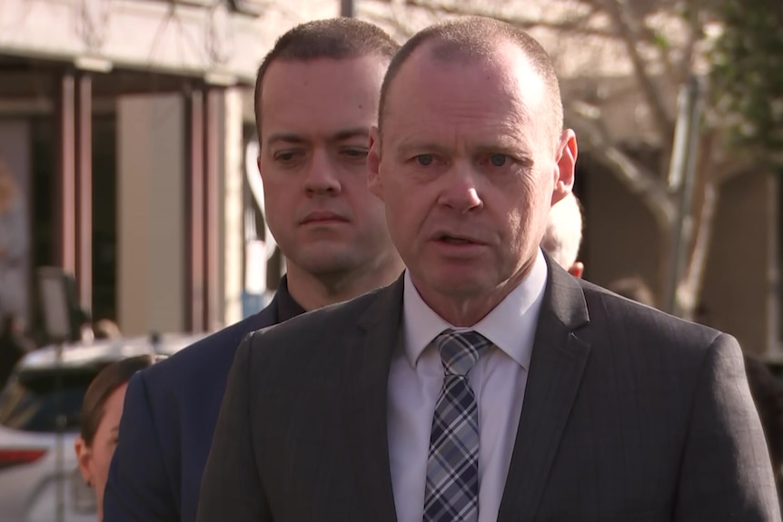 A man talking to the media, with another man in the background