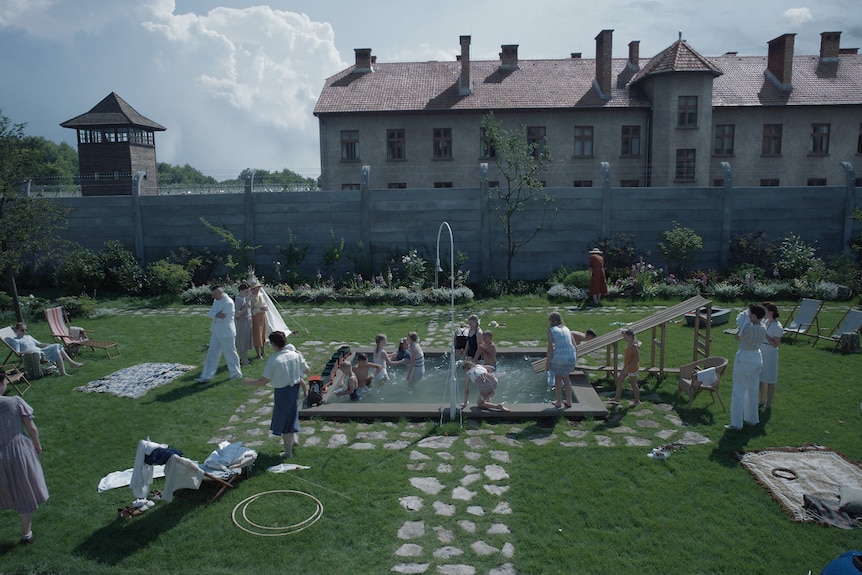A film still of a garden party. The people are in 40s-style dress. Behind them one of the buildings of Auschwitz looms.