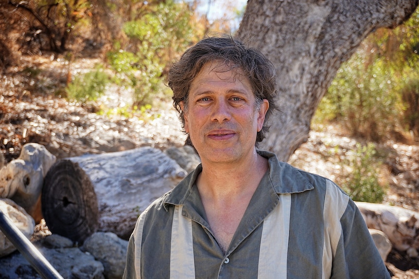 A man wearing a green-ish button up shirt with thick white stripes standing in front of trees, staring into camera