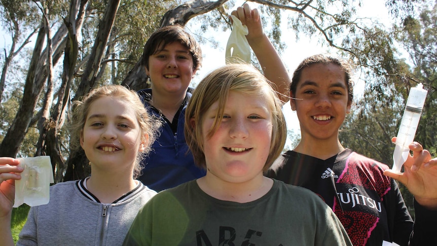 Four children holding a syringe, rubber gloves and a small plastic tube.