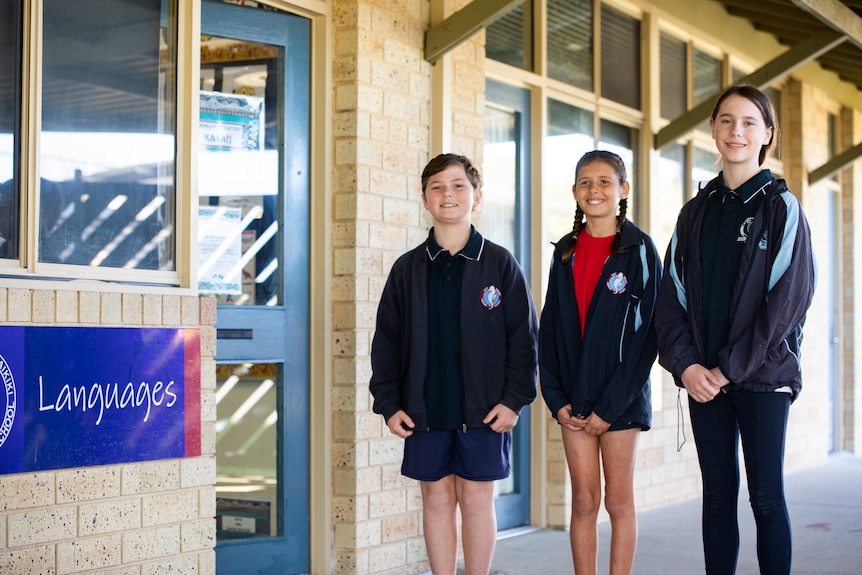 Three children stand outside a classroom smiling at the camera.