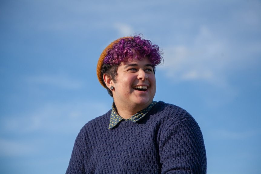 A man with purple hair looks towards the right, laughing. The camera is at a low angle, meaning the blue sky is the background