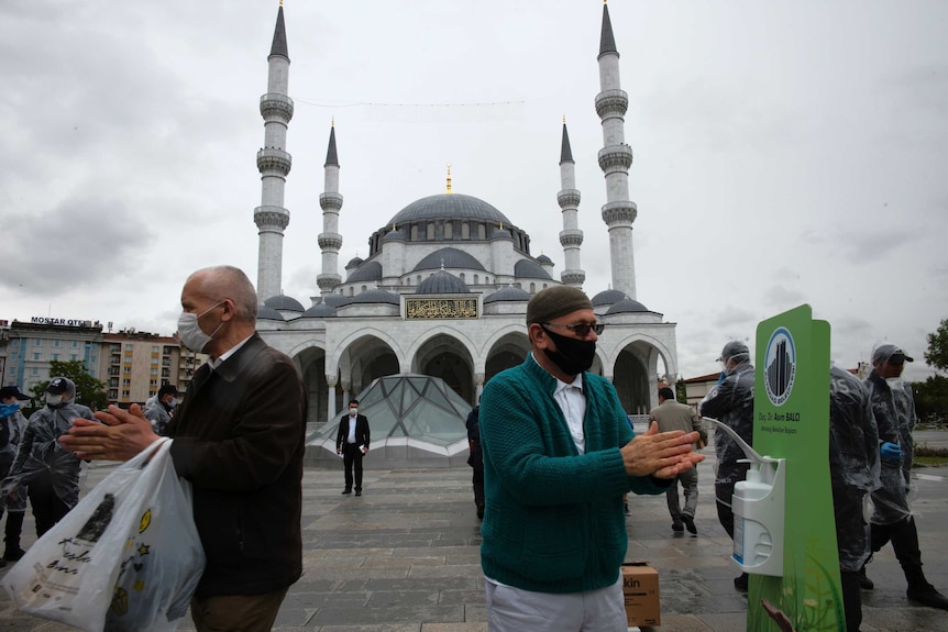 Two middle aged men wearing face masks sanitise their hands as the stand in the forecourt of a large blue and white mosque.