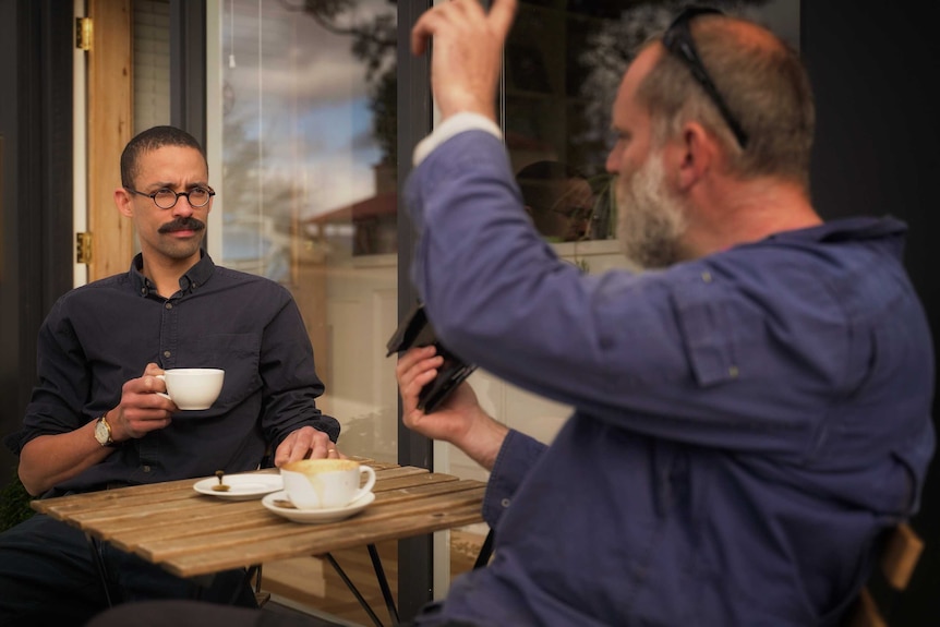 Two middle aged men sit at an outdoor table drinking coffee