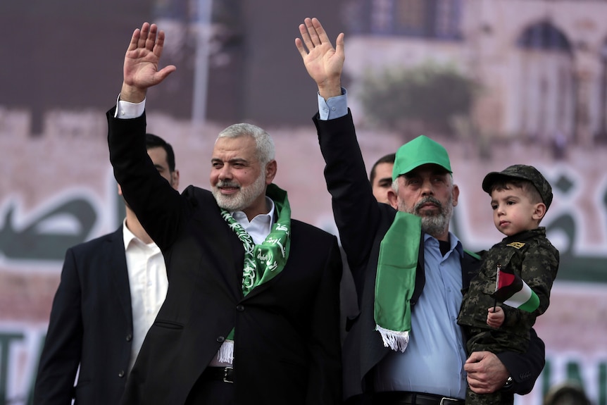 A man wearing a green cap and holding a child waves to a crowd beside another man dressed in a suit and scarf.