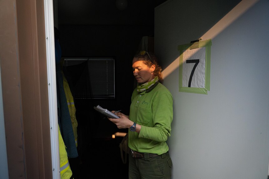 A woman in a green shirt stands in the doorway of a demountable, looking at a notebook. A ray of light shines across her face.