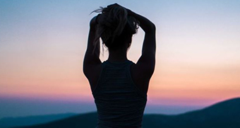 Silhouette of a woman standing with hands above her head facing a sunset and mountains in the distance.