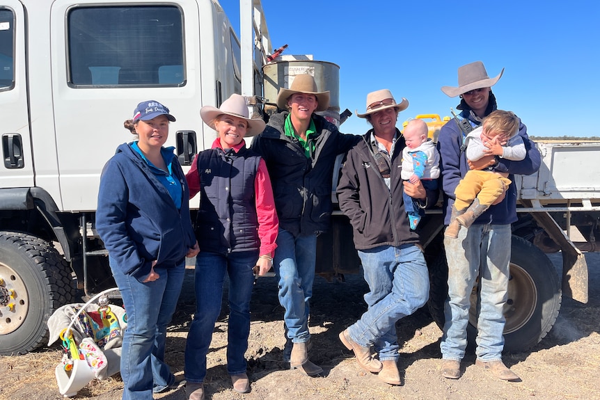 Two women, three men and two children stand next to a large truck wearing outdoor clothing and cowboy hats. 