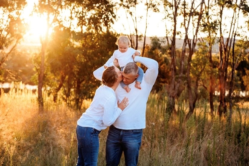 A man and a woman stand with a young child on the father's shoulder, in a field at sunset.