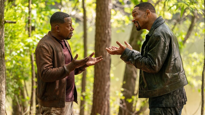 Two men in leather jackets stand in a forest. They are facing each other, both standing with arms up in a confused shrug.