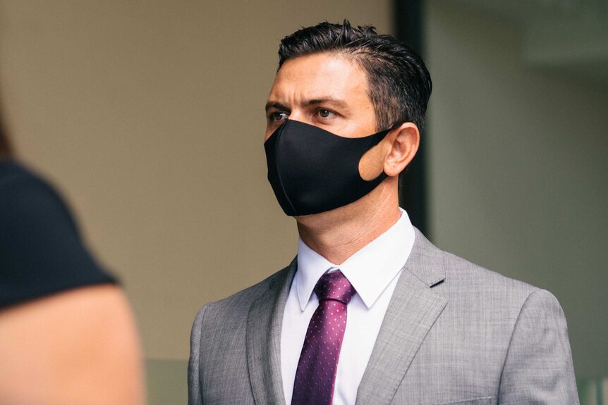 A close up of Nathan wearing a grey suit and purple tie, with a black face mask, walking into court.