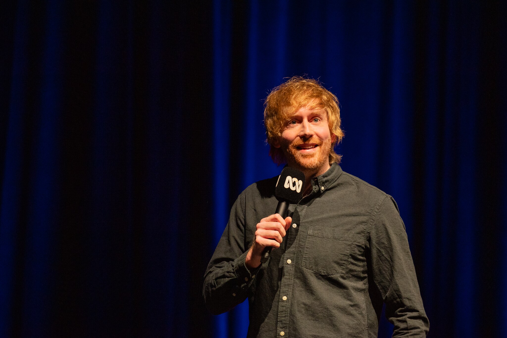 Mark Simmons on stage with dark blue stage curtains in the background.