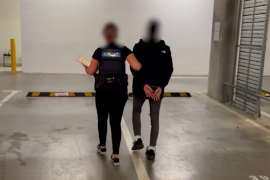 A teenager is escorted in a car park by a female officer. Their faces are blurred.