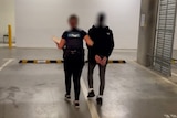 A teenager is escorted in a car park by a female officer. Their faces are blurred.