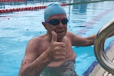Elderly man giving thumbs up in pool with swimming cap.