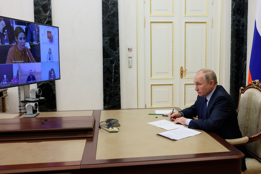 Vladimir Putin sits with pen in hand as he views TV screen across table of videoconference. 
