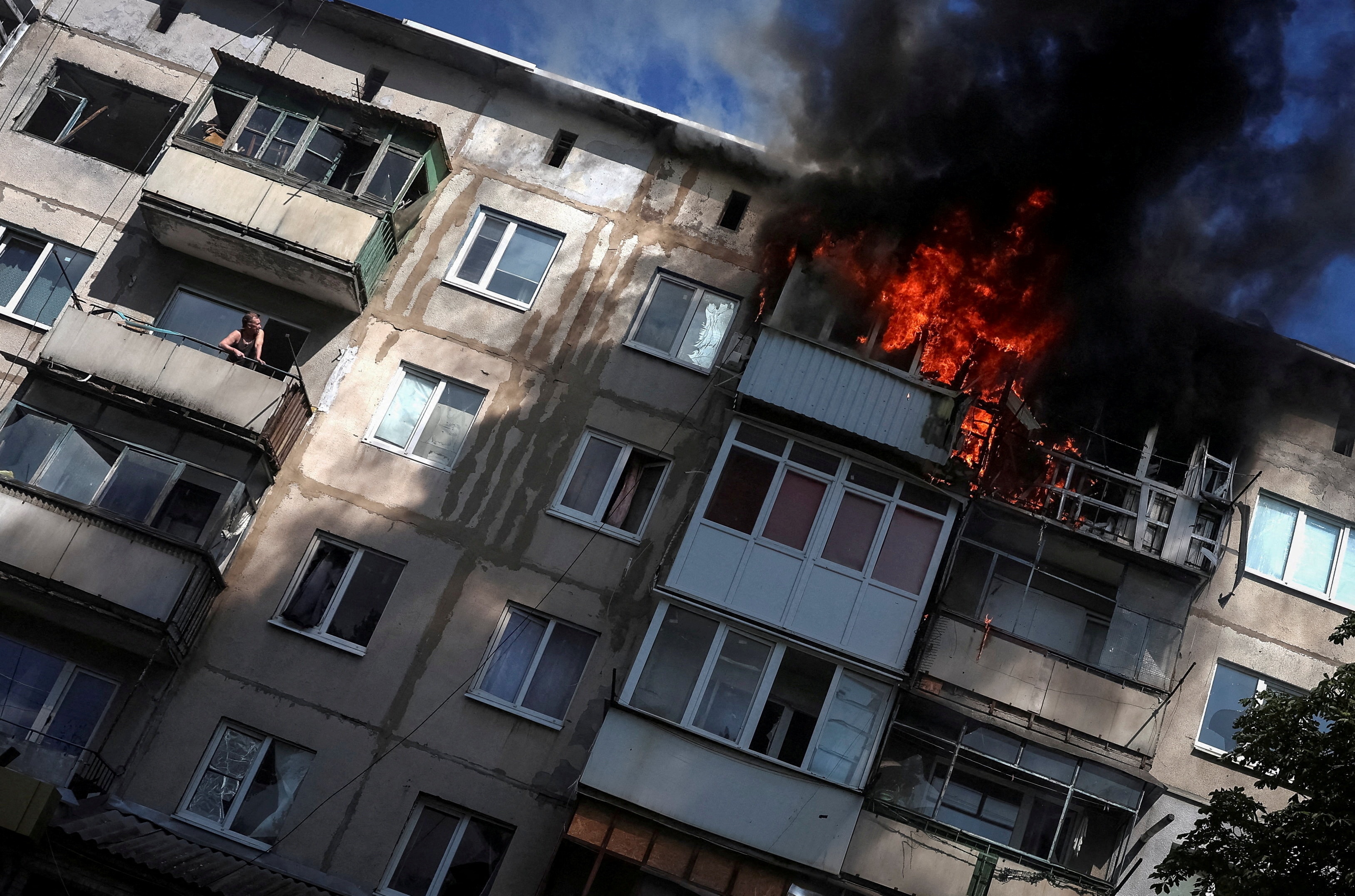 A window from an apartment building has blown open and flames are pouring out of it.