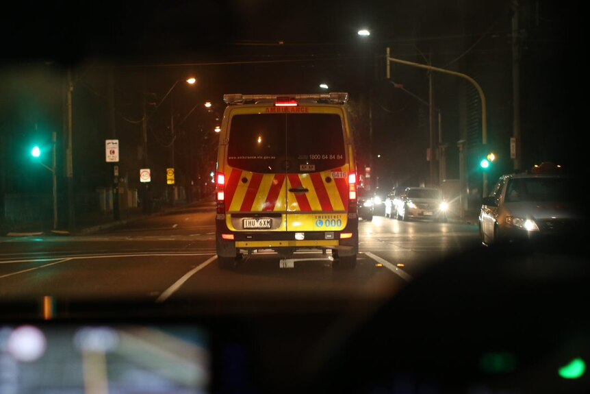 An ambulance can be seen through the windscreen of the car behind, driving along a Melbourne road at night.