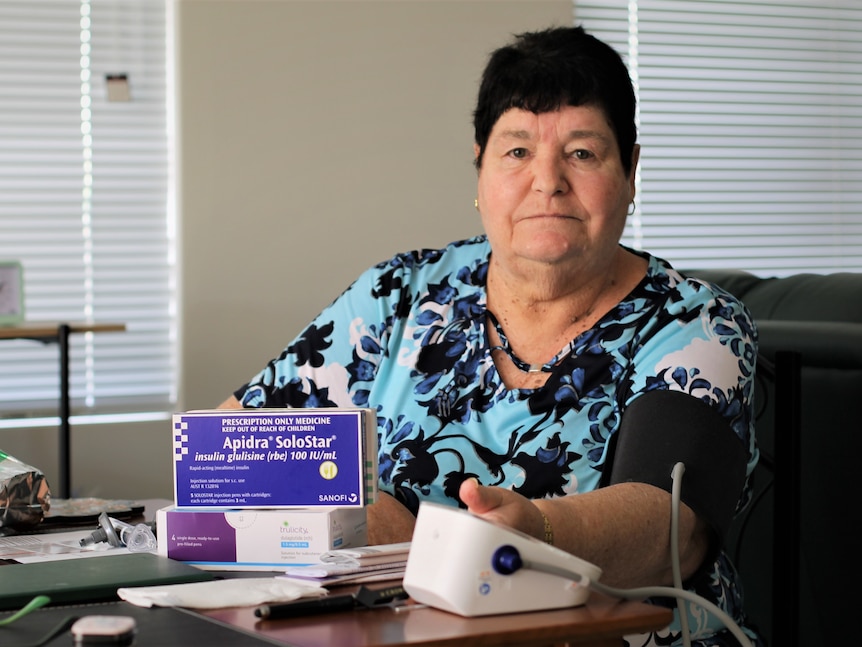 Jennifer wears a blue floral print top and sits at a table with boxes of her medication