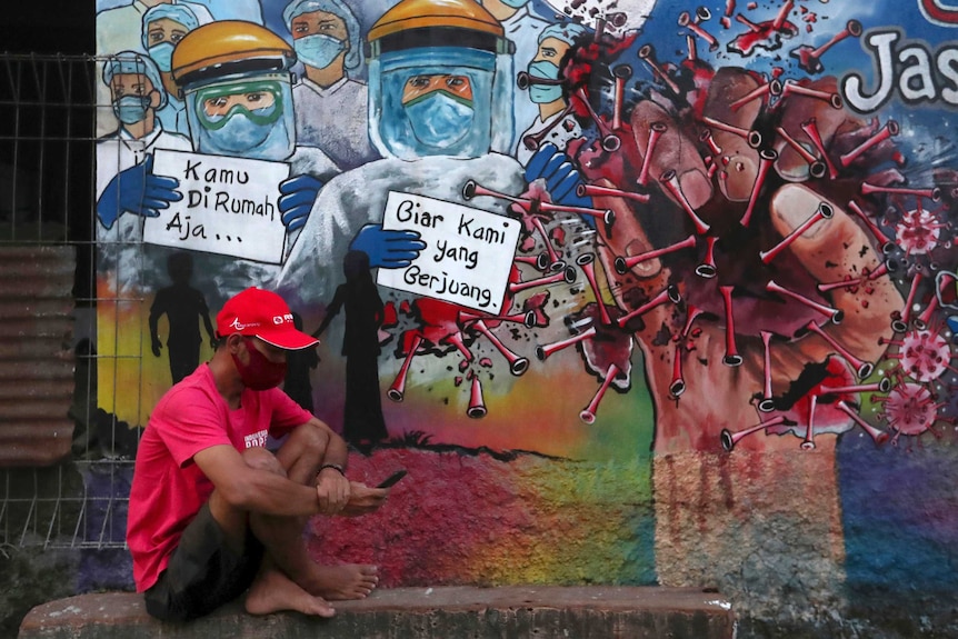 A man sits on the street in front of a coronavirus-inspired mural which reads: "you just stay home, to let us fight."