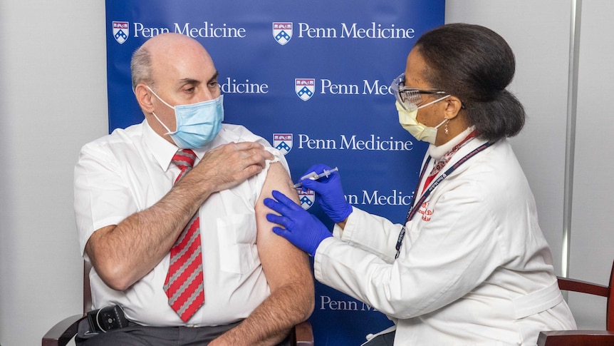 Drew Weissman is injected with a COVID-19 vaccine. He is wearing a mask and looks serious.