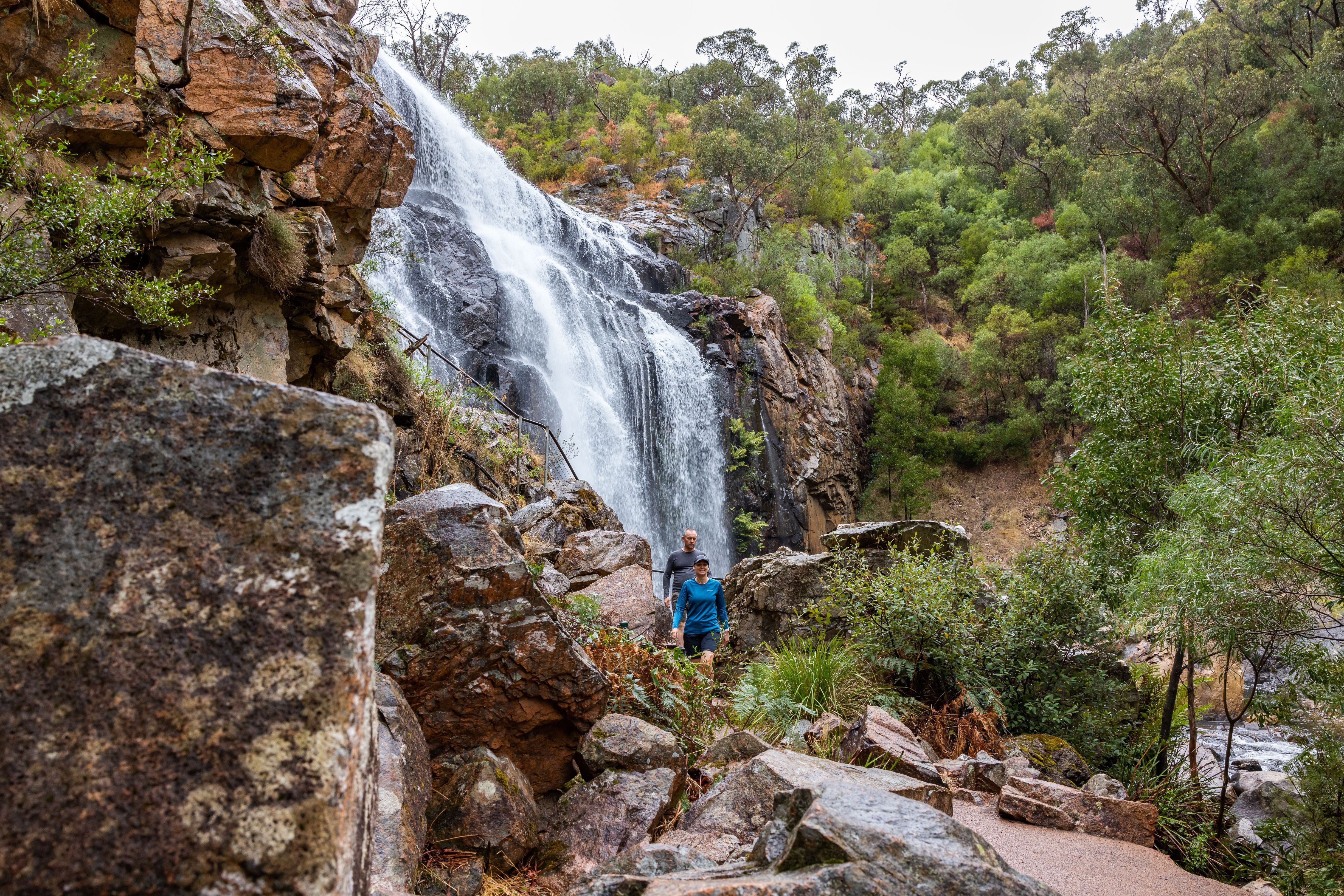 Two hikers in front of a waterfall among rocks and trees.
