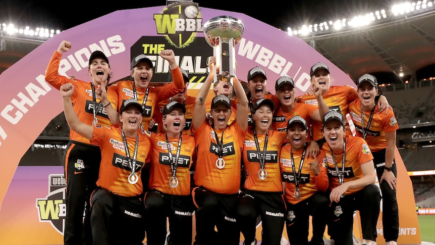 A group of women cricketers in orange hold a trophy aloft and shout in joy in middle of stadium