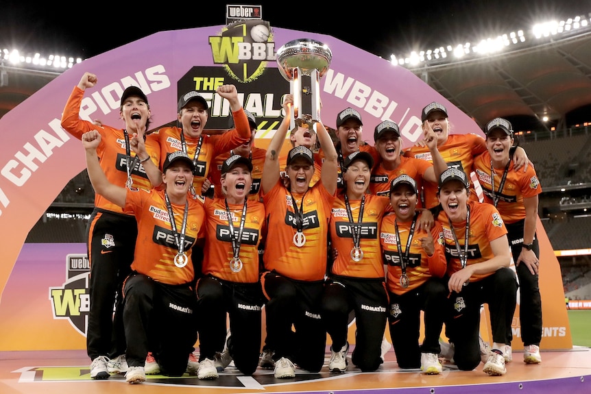 Un grupo de jugadoras de críquet vestidas de naranja sostienen un trofeo en alto y gritan de alegría en medio del estadio