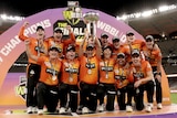 A group of women cricketers in orange hold a trophy aloft and shout in joy in middle of stadium