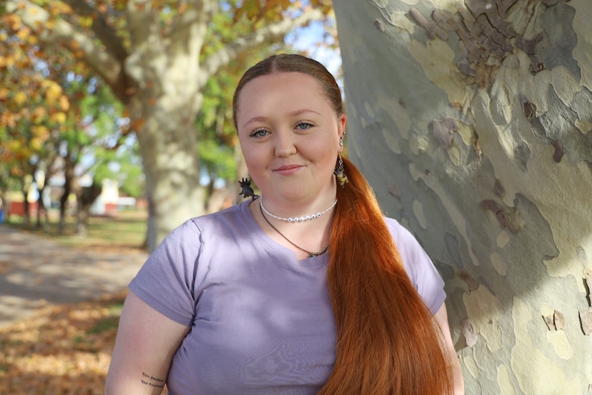 A woman with long, red hair and wearing a purple T-shirt smiles at the camera as she stands next to a tree