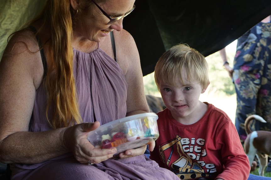 Woman in purple dress holding tub of lollies, sitting inside tent with young boy in red shirt