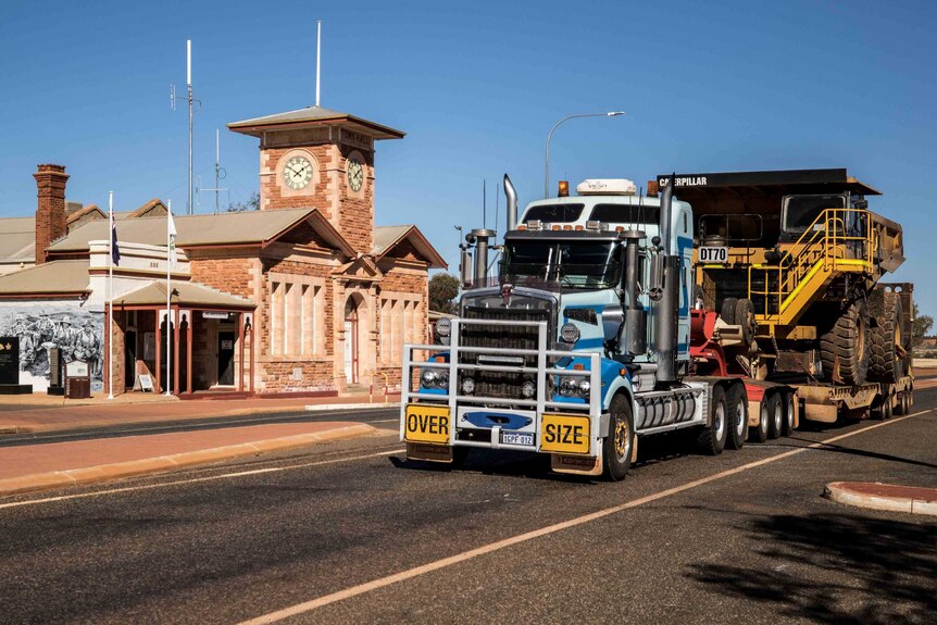 A road train carrying a mining truck drives through a rural townsite.