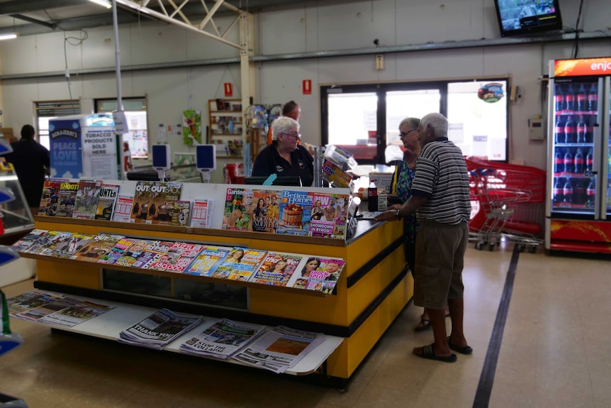 A woman serves an elderly man and woman from behind a yellow checkout stacked with magazines.