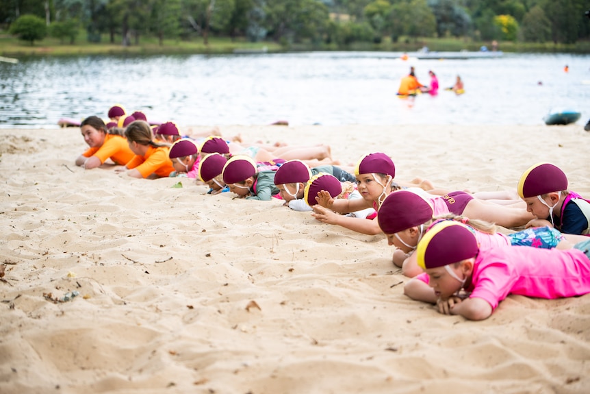 Several kids in swimming caps and pink shirts lie in a line on the sand by a lake.