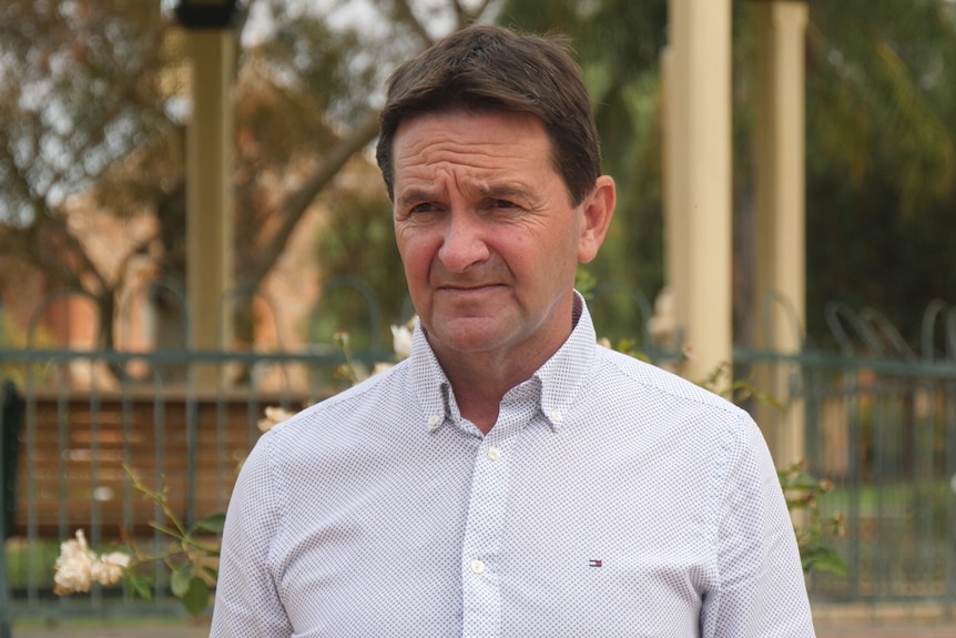 A white man with brown hair wearing a white patterned shirt standing in a park.