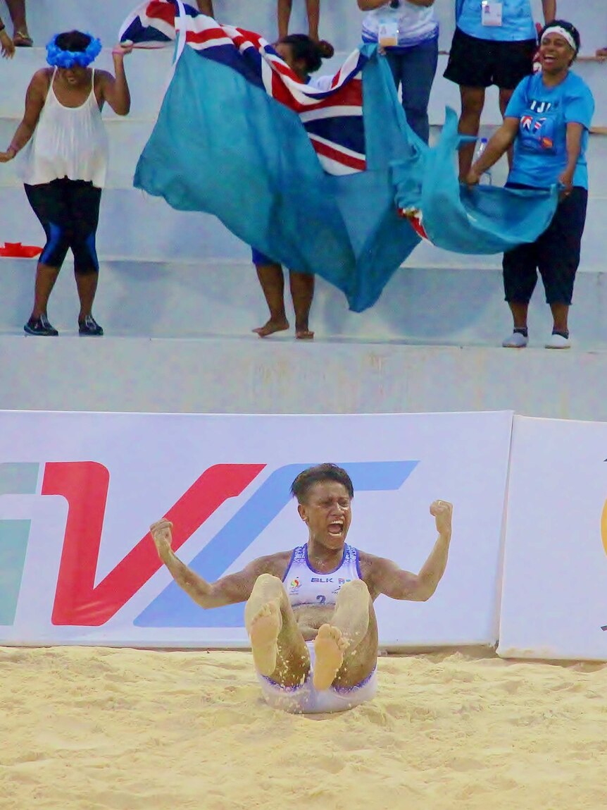 A Fijian woman in a beach volleyball uniform sits in the sand and pumps her fists.