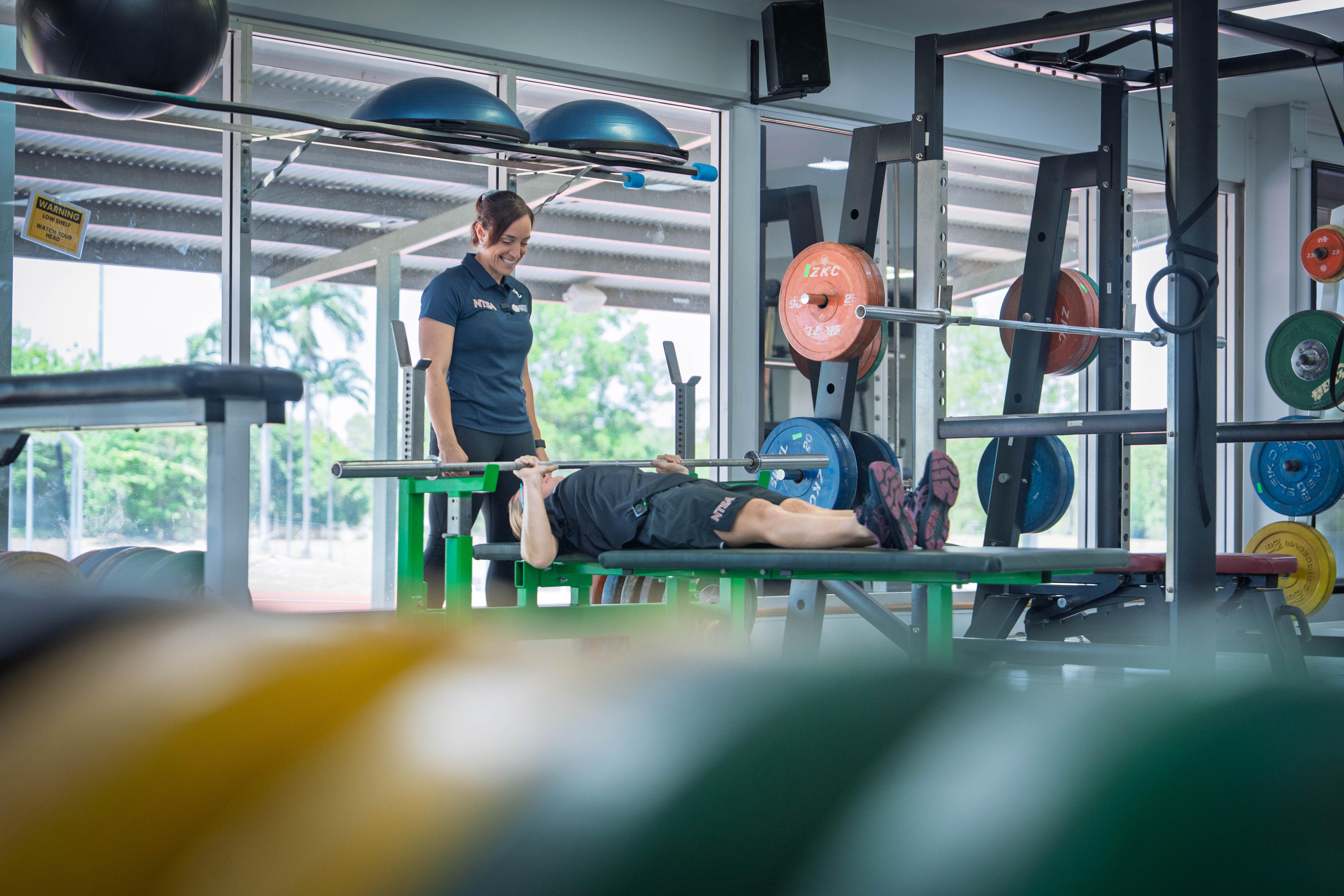 Toma de gran angular de una mujer blanca tumbada sobre un equipo de fitness y haciendo ejercicios con barra mientras el entrenador está encima de ella.