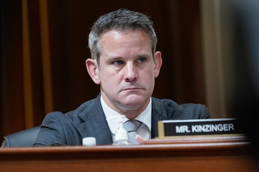 adam kinzinger sits at a desk behind a nameplate with his surname on it with a serious look on his face